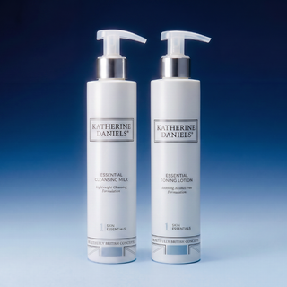 Two white pump bottles of Katherine Daniels skincare products, Essential Cleansing Milk and Essential Toning Lotion, standing side by side on a blue gradient background.