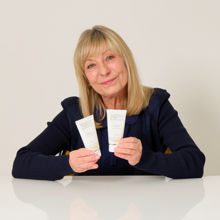 A smiling blonde woman seated at a white table holds up two white skincare tubes, one in each hand, against a plain light beige background.