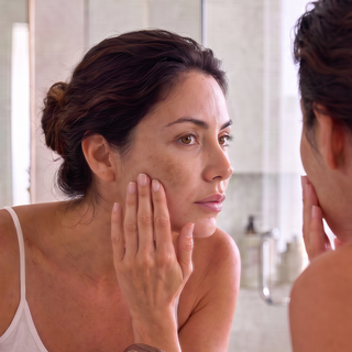 A woman looking at her reflection in a bathroom mirror, touching her face. She is wearing a white spaghetti-strap tank top, and the background shows a softly blurred bathroom setting with bottles on a counter.