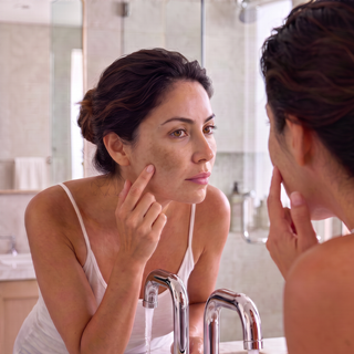 A woman in a white camisole looks at her reflection in a bathroom mirror, touching her cheek as if checking skin texture or makeup. Chrome faucets and a tiled bathroom are visible in the background.