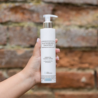 Hand holding a bottle of Katherine Daniels essential toning lotion against a brick wall.